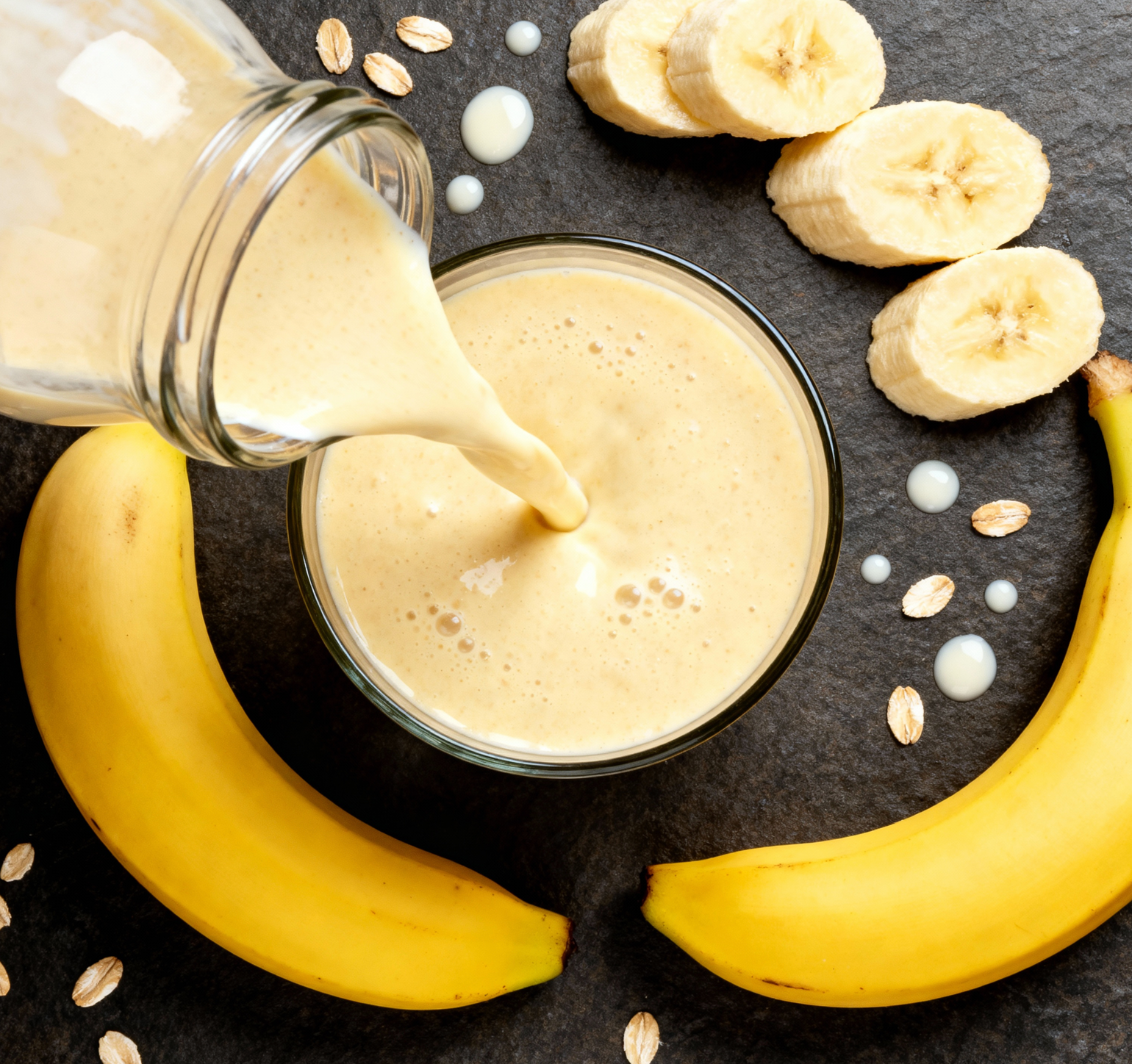 Banana smoothie being poured into a glass with bananas and oats on a dark surface